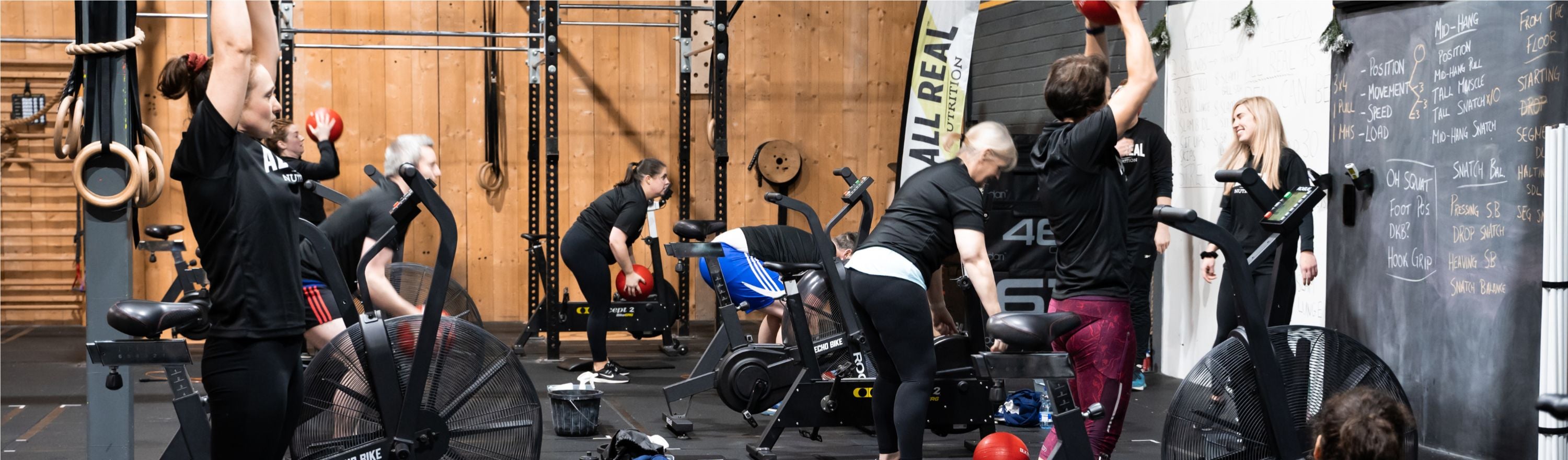 Group fitness class in a gym with people exercising on bikes and lifting medicine balls, All Real Nutrition banner visible