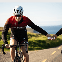 Cyclist in red and black uniform receiving an All Real Peanut Butter Protein Bar from another person on a road with scenic background.