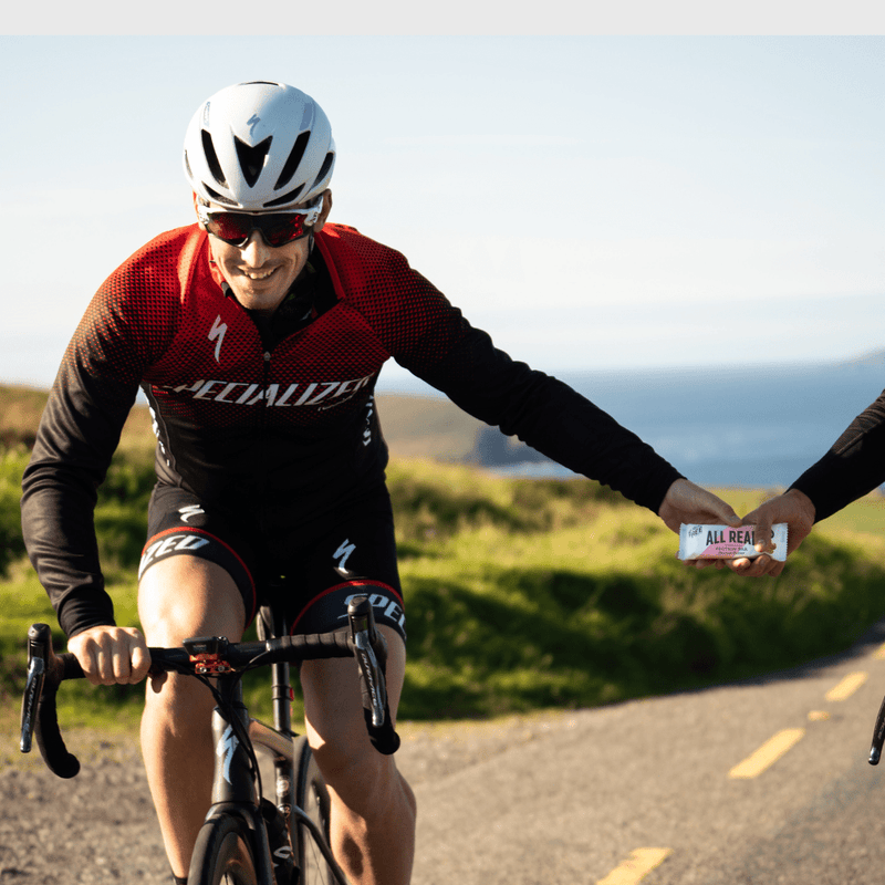 Cyclist in red and black uniform receiving an All Real Peanut Butter Protein Bar from another person on a road with scenic background.