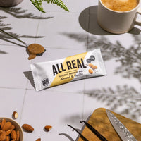 All Real almond vanilla protein bar with almonds, vanilla pods and coffee on table