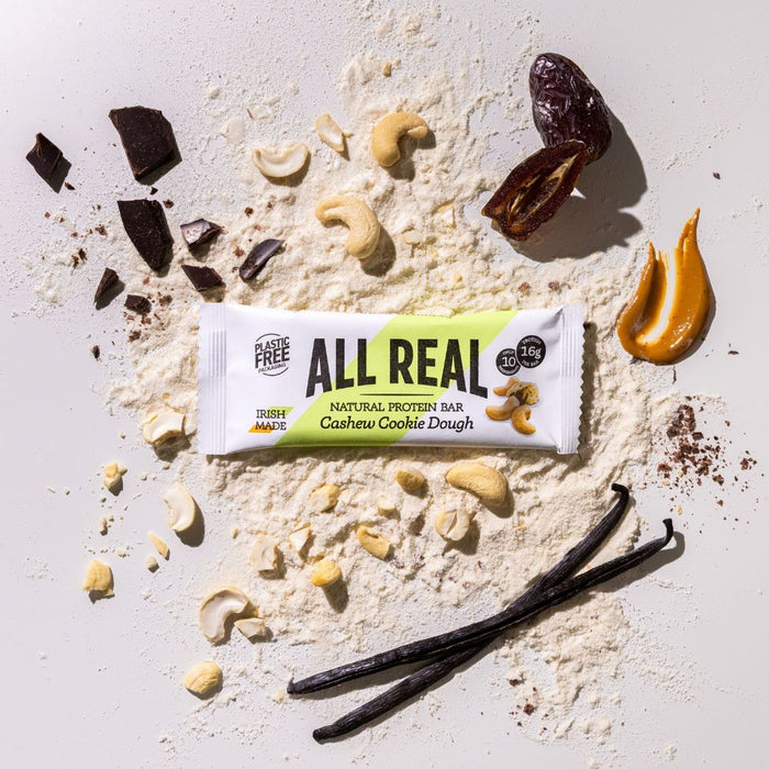 All Real cashew cookie dough protein bar with cashews and cookie dough ingredients scattered around