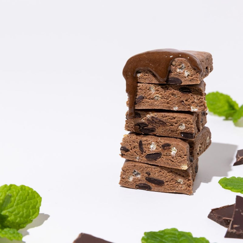 All Real chocolate mint protein bar pieces stacked with melted chocolate drizzle, mint leaves and chocolate chunks on white background