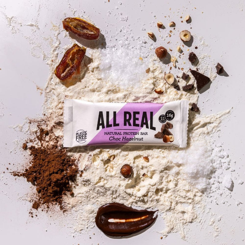 All Real chocolate hazelnut protein bar with hazelnuts and chocolate pieces on ingredient scatter background