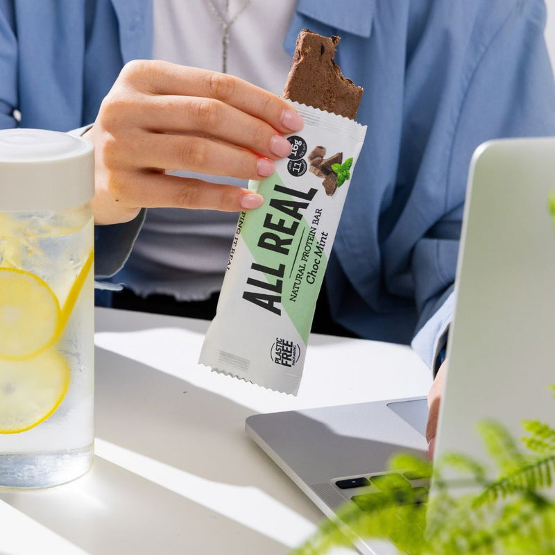 Person holding All Real chocolate mint protein bar beside laptop