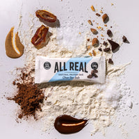 All Real chocolate sea salt protein bar with cocoa powder and chocolate pieces on ingredient scatter