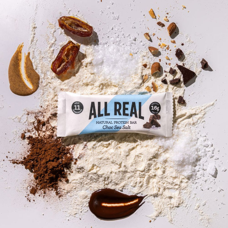All Real chocolate sea salt protein bar with cocoa powder and chocolate pieces on ingredient scatter