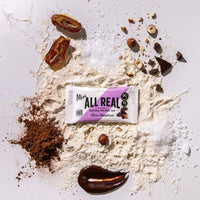 Mini All Real chocolate hazelnut protein bar with hazelnuts and chocolate ingredients scattered around