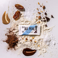 Mini All Real chocolate sea salt protein bar with cocoa and chocolate ingredients scattered around