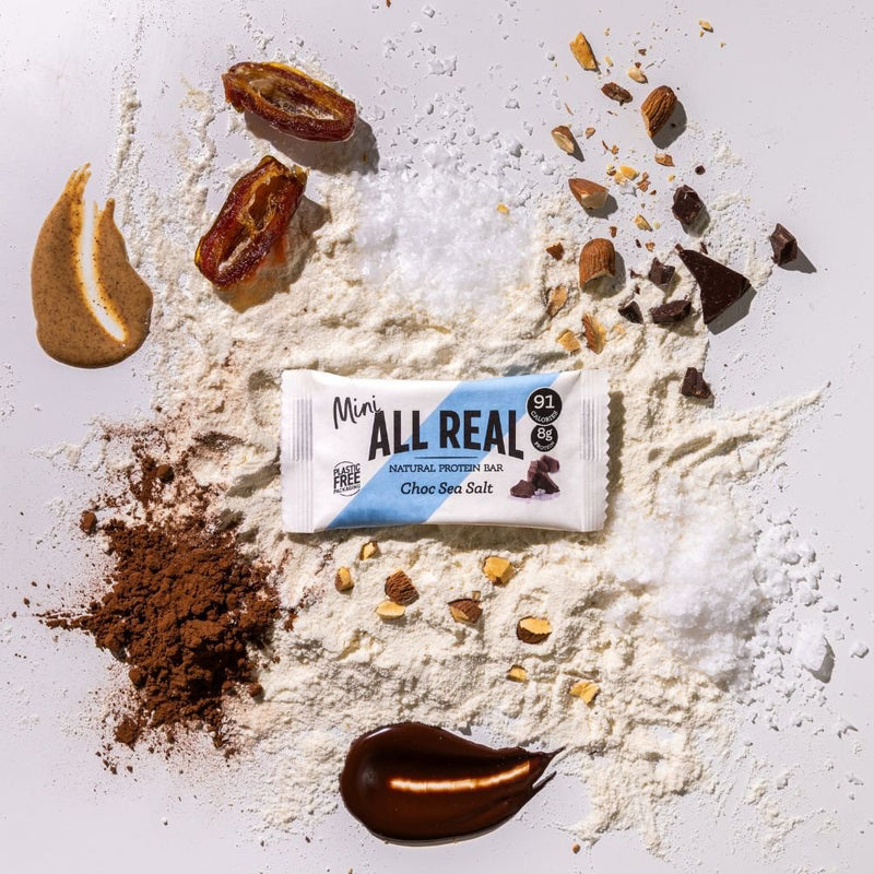 Mini All Real chocolate sea salt protein bar with cocoa and chocolate ingredients scattered around