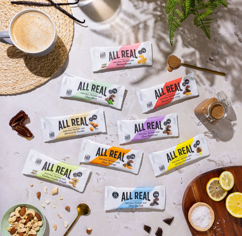 Variety of All Real natural protein bars displayed in a flat lay with coffee, nuts, chocolate and fruit ingredients showing the full range of Irish-made high protein snack bars.