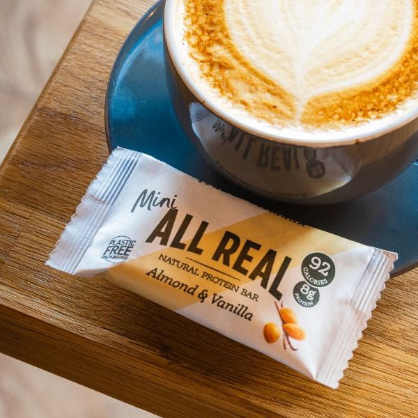 All Real almond and vanilla protein bar next to a cup of coffee on a wooden surface